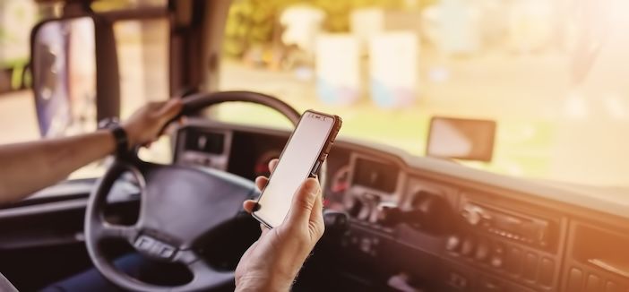 A driver’s hand holds a smartphone in front of the dashboard of a truck cab while the other hand rests on the steering wheel.