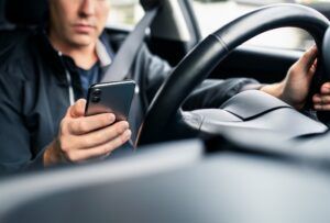 A man in a dark jacket wearing a seatbelt looks down at a smartphone in one hand while his other hand rests on the steering wheel of a car.