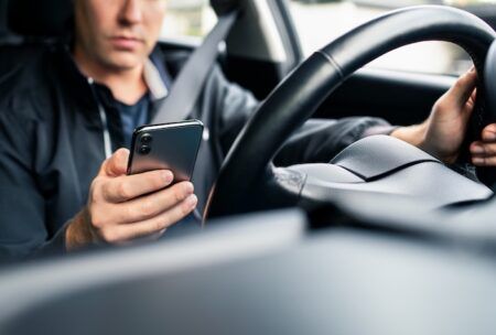 Distracted driving tech showcased at US safety event A man in a dark jacket wearing a seatbelt looks down at a smartphone in one hand while his other hand rests on the steering wheel of a car.