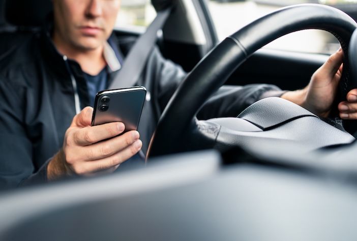 A man in a dark jacket wearing a seatbelt looks down at a smartphone in one hand while his other hand rests on the steering wheel of a car.