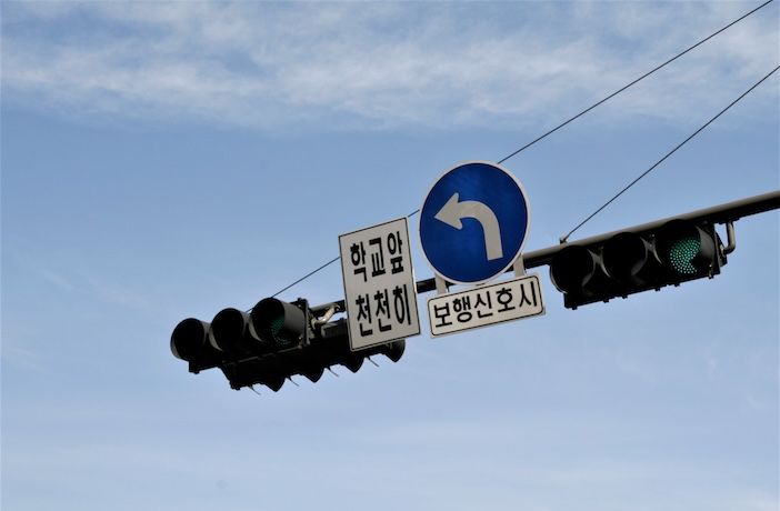 Traffic signals on a gantry arm against a blue sky, with green lights lit, a blue left-turn arrow sign and two Korean-language signs below