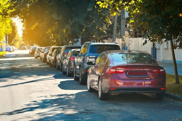 A row of cars parked along a tree-lined street in low sunlight, with a maroon saloon in the foreground.
