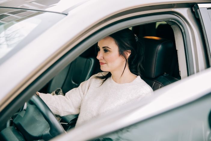 A woman with dark hair tied back, wearing a cream knitted jumper, sits in the driver's seat of a car and looks ahead, seen through the open side window.