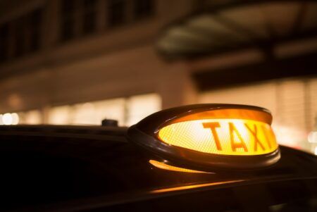 London autonomous transport report: GMB urges driver plan Close-up of an illuminated amber taxi roof sign glowing at night on a dark vehicle, with blurred lit buildings behind.