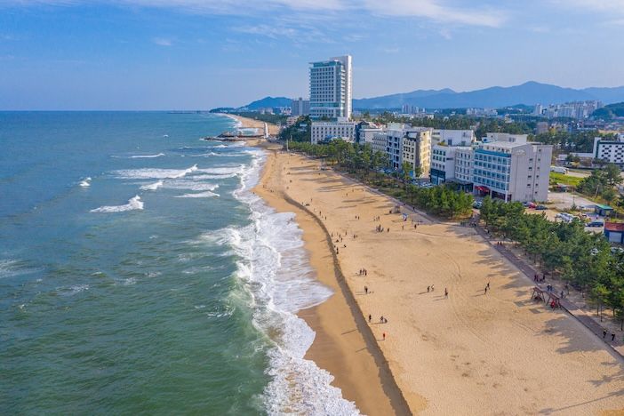 Aerial view of a wide sandy beach in Gangneung, with waves breaking on the shore and hotels and mountains behind