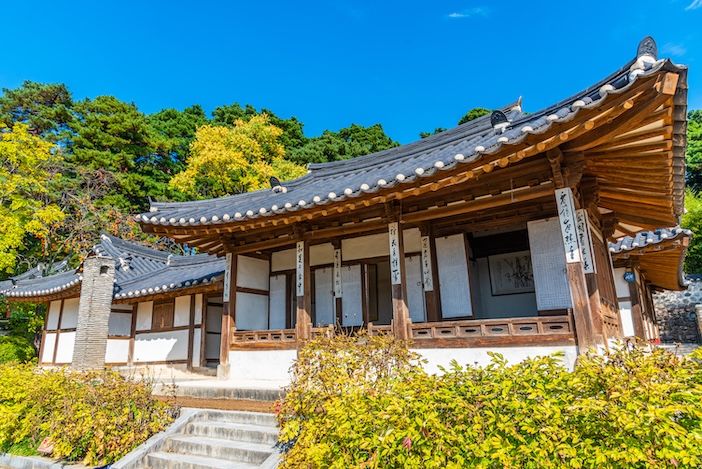 Traditional Korean wooden house with a tiled roof and open veranda, framed by trees in autumn colour