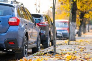 A blue crossover and other cars parked beside a pavement covered in yellow autumn leaves.