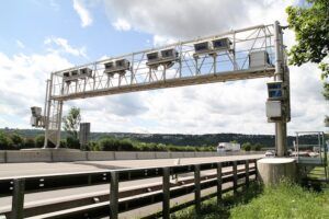 A white steel gantry spanning a motorway with a row of boxed cameras mounted along it, beside a crash barrier, under a partly cloudy sky with a lorry in the distance