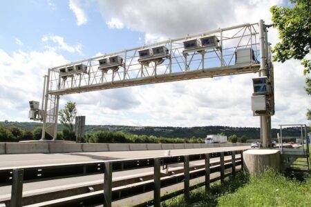 A white steel gantry spanning a motorway with a row of boxed cameras mounted along it, beside a crash barrier, under a partly cloudy sky with a lorry in the distance