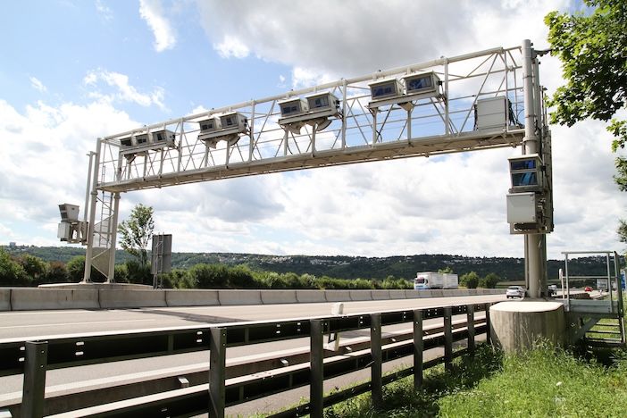 A white steel gantry spanning a motorway with a row of boxed cameras mounted along it, beside a crash barrier, under a partly cloudy sky with a lorry in the distance