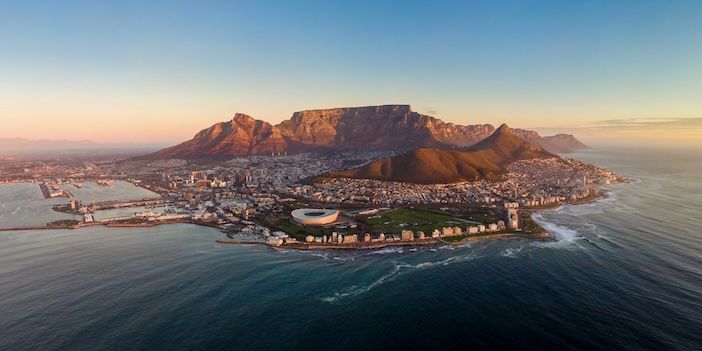 Aerial view at sunset of a coastal city beneath a flat-topped mountain, with a stadium beside the sea.