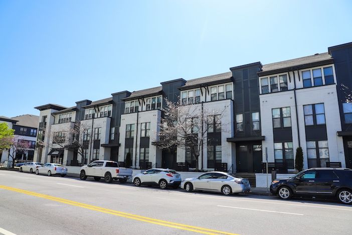 Cars parked at the kerb in front of a row of white and black multi-storey townhouses under a clear blue sky.