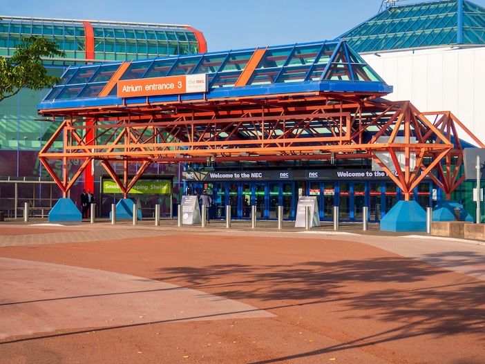 An orange and blue steel-framed canopy over a glass-fronted entrance, with a paved forecourt in the foreground and a clear blue sky above.
