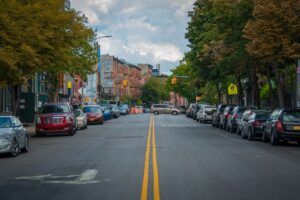 A tree-lined city street with cars parked along both kerbs, brick buildings in the distance and a yellow pedestrian crossing sign on the right.