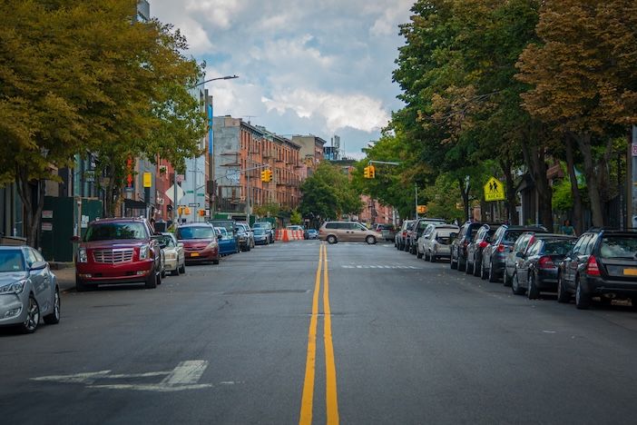 Arrive completes Passport acquisition for kerb management A tree-lined city street with cars parked along both kerbs, brick buildings in the distance and a yellow pedestrian crossing sign on the right.