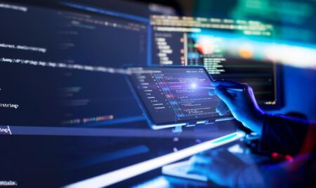 HIGHWAYS UK: Updated highway infrastructure code of practice published A hand holds a stylus to a tablet showing lines of code, in front of large monitors displaying code in a dark, blue-lit room.