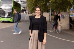 A smiling woman in a black blazer and beige wide-leg trousers stands on a city street, with a green double-decker bus, a pedestrian and a cyclist blurred behind her.