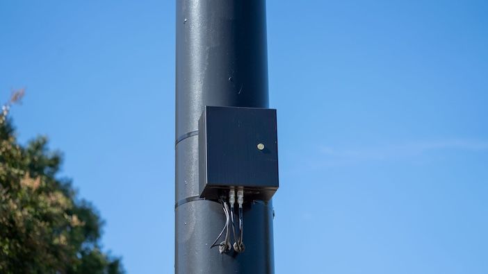 square black box fixed to a dark pole, with cables running out of the bottom, against a blue sky.