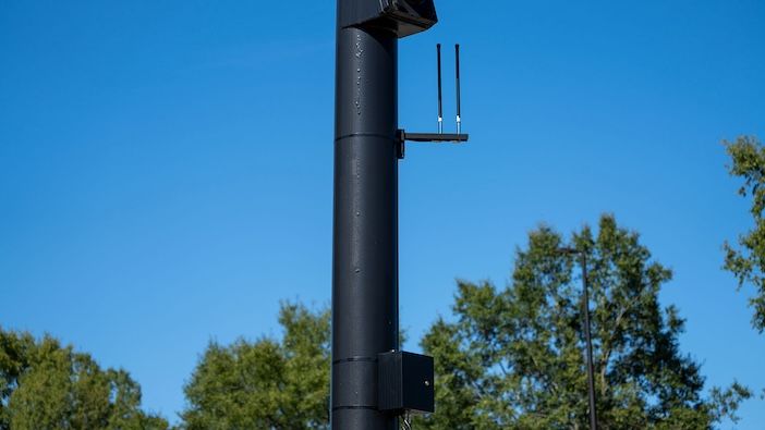 Applied Information C-V2X roadside unit gains OmniAir certification A tall black pole with a pair of antennas on a bracket near the top and a black box lower down, with trees behind.