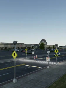 Illuminated pedestrian crossing signs positioned either side of a marked crosswalk at a retail park, with two people crossing the road at dusk.