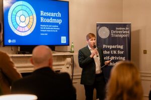 A man in a dark green jacket speaks into a microphone beside a wall-mounted screen showing a colourful circular chart and the words Research Roadmap, with a navy banner behind him.