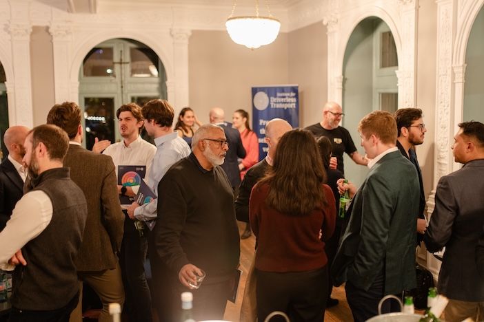 uests stand talking in a high-ceilinged room with arched windows, some holding printed booklets and drinks, with a navy banner in the background.