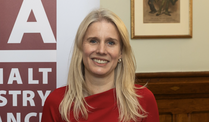 A woman with long blonde hair and a red dress smiles while holding a printed report, standing in front of a banner in a wood-panelled room.