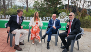 Four people sit on grey folding chairs outdoors in conversation, one holding a microphone, in front of a large green ITS World Congress 2026 sign and trees