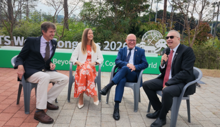 PODCAST 59: Live at the ITS World Congress for ERTICO’s 35th Four people sit on grey folding chairs outdoors in conversation, one holding a microphone, in front of a large green ITS World Congress 2026 sign and trees