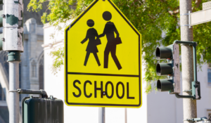 Yellow warning road sign for a school zone at a road intersection with a street traffic light in the background