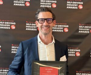A smiling man in a navy suit and glasses holds a framed certificate in front of a dark backdrop repeating a self-driving awards logo.