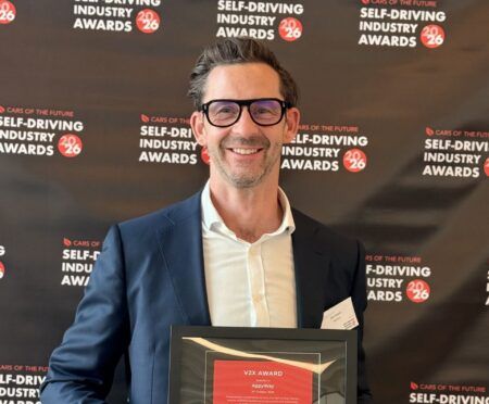 AppyWay wins V2X Award at Self-Driving Industry Awards 2026 A smiling man in a navy suit and glasses holds a framed certificate in front of a dark backdrop repeating a self-driving awards logo.