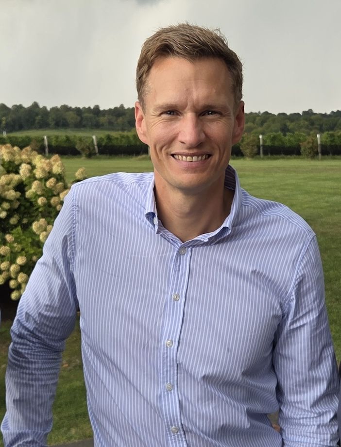 A smiling man in a light blue striped shirt stands outdoors in front of a flowering bush, a lawn and a line of trees.