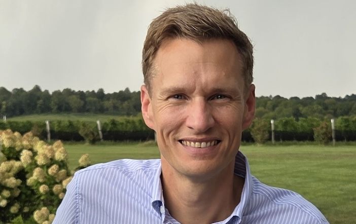 A smiling man in a light blue striped shirt stands outdoors in front of a flowering bush, a lawn and a line of trees.