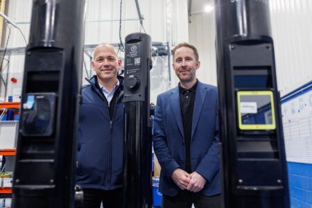 Connected Kerb opens Aberdeen EV charger manufacturing hub Two men stand either side of a black charging post in a workshop, with more charging posts in the foreground and a whiteboard on the wall behind them.