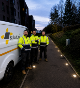 Three men in high-visibility jackets stand beside a white van on a footpath at dusk, with a line of small lit studs set into the tarmac beside them.