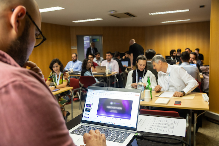A group of people seated at tables in a wood-panelled room, with a laptop showing a slide in the foreground and a bearded man in glasses looking on.