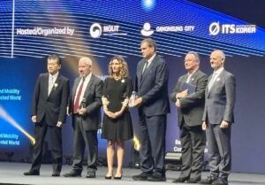Six people stand in a line on a stage in front of a lit blue screen. From the left: a man in a dark suit with a striped tie and a badge on his lapel; an older man with white hair and a beard in a dark suit and red tie, holding a small trophy; a smiling woman with curly hair in a black dress with lace sleeves; a tall man in a navy suit holding a glass trophy; a man with glasses in a grey jacket holding a dark folder; and a man with a short grey beard in a grey suit and light-coloured tie. A banner above them reads ‘Hosted/Organized by’ beside three organisation logos.